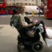 Hannah sitting in a trial powerchair (wheelchair), outside of hospital for the first time in several years. Warren Street tube station is in the background.