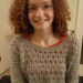 Hannah sitting in her powerchair wearing a grey jumper with holes in it and an orange long sleeve t-shirt underneath. She is smiling at the camera. She has thick curly aburn hair and is wearing glasses.
