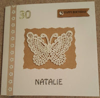 A white card with beige ribbon with white flowers on it down one side. A white lace butterfly in the middle on top of a brown square of paper. A silver 30 on the top left next to the ribbon and the name 'Natalie' at the bottom in the middle. There is also a brown sign on the top right hand corner with the words 'Happy Birthday' in black writing on it.