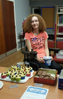 Hannah sitting in her powerchair, holding a pot of money. In front of her on the table are malteaser cupcakes and malteaser traybake. Hannah is smiling at the camera.