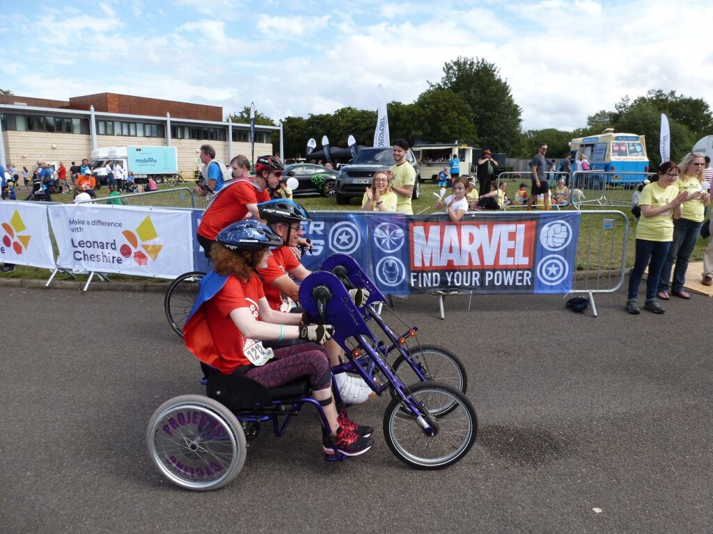 Hannah and Brian handcycling at the Superhero Tri. They are in a side by side upright handcycle, wearing helmets. Matt is behind cycling on a standard bike.