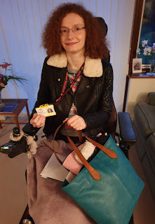 Hannah after her first day of work experience Hannah, a young woman with curly aburn hair and glasses, wearing a leather jacket and sitting in her powerchair. She is holding a turquoise handbag in her left hand and holding up her work badge in her right hand.