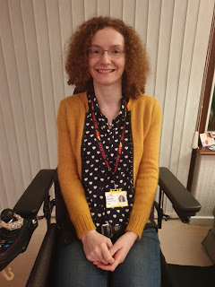 Hannah after work Hannah- a young woman with curly aburn hair and glasses sitting in her powerchair. She is wearing jeggings, a blue blouse with white hearts and a mustard cardigan