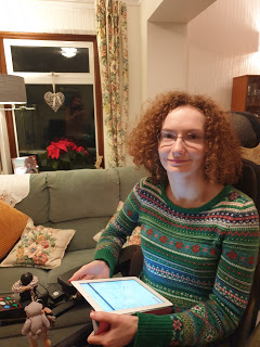 Hannah-a young woman with curly aburn hair and glasses, wearing a green patterned jumper and holding her iPad.