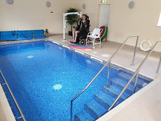 The swimming pool and hoist with Hannah sitting in her powerchair with her hoist sling under her and attached to the hoist. She is about to be hoisted into the pool.