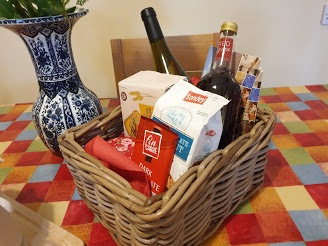A straw box with several bottles of drink, biscuits, chocolate and homemade jam in it.