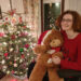 Hannah sitting in her powerchair wearing a red minnie mouse christmas jumper. She has her big Christmas bear-Dandy in her arms and the Christmas tree is next to her decorated.