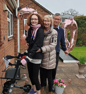 Hannah with her parents, after completing stage 1 of her #BenHannah3530 challenge. There is a big 3 and 0 balloon behind them in rose gold. They are smiling at the camera.