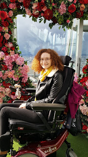 Hannah sitting in her powerchair, she is wearing a leather jacket and smiling at the camera. She is surrounded by pink and red flowers and the city of London is in the background. She has been nominated for a National Diversity Award.