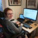 Hannah sitting in her powerchair under a desk with a laptop, monitor, keyboard and mouse on it. She is smiling at the camera.