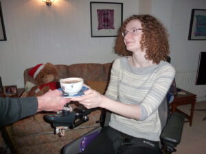 Hannah sitting in her powerchair smiling at one of her PAs who is giving her a hot chocolate.