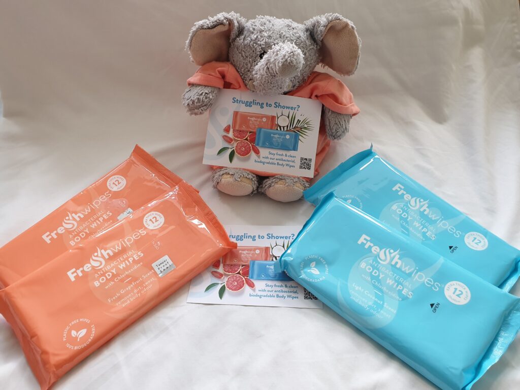 Nellie my elephant holding a FreshWipes leaflet which says: 'Struggling to shower' and has a picture of the grapefruit and coconut FreshWipes on it. Next to her are two orange packets of grapefruit and then two blue packets of coconut FreshWipes.