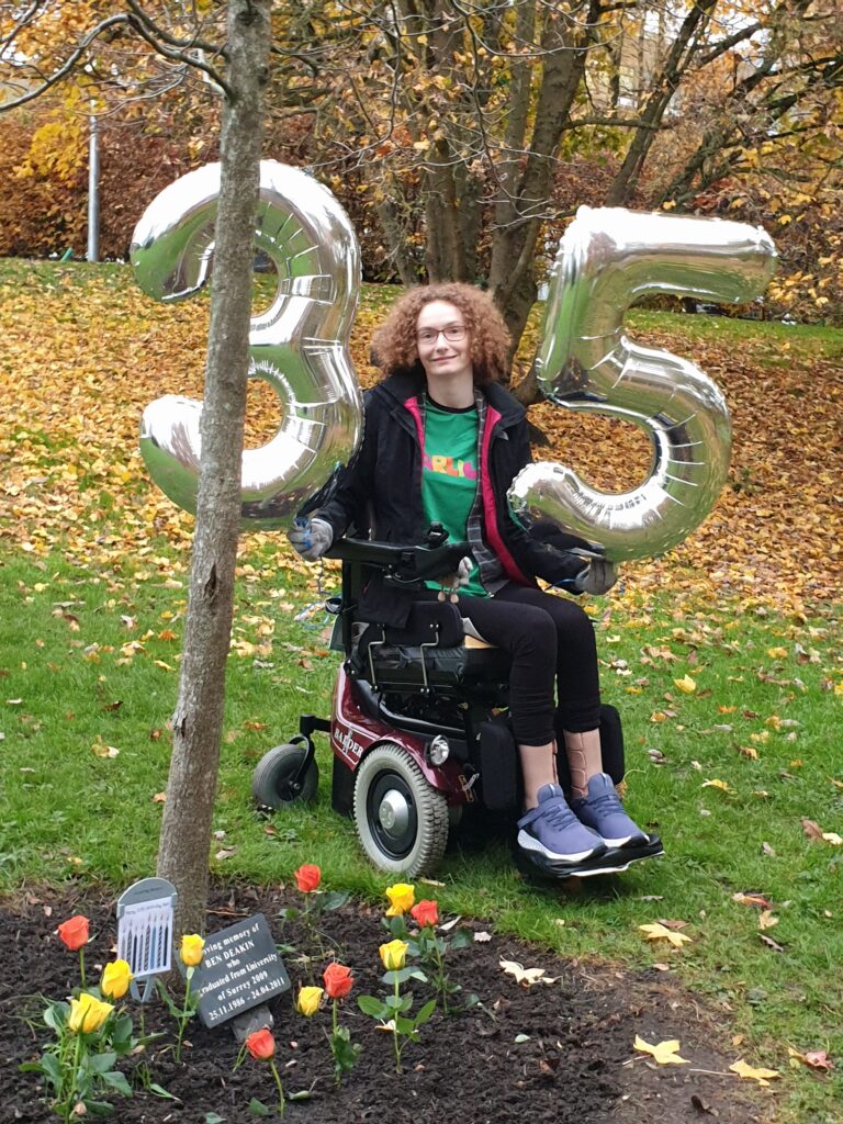 Hannah is sitting in her powerchair after completing the final length of her #BenHannah3530 challlenge. She is holding a silver 3 and 5 number balloon and sitting next to Ben's tree. There is a slate plaque with Ben's name on at the bottom of his tree, a birthday card and yellow and orange roses.