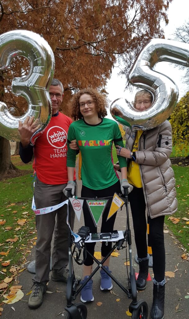 Hannah stepping through the finish line with her four wheeled walker and her parents supporting her each side. She is wearing a green Starlight charty t-shirt, black leggings, splints and purple trainers. Her Dad, Brian is wearing a red White Lodge charity t-shirt and holding a silver 3 number balloon. Her Mum, Jill is wearing a yellow Child Bereavement UK t-shirt and holding a 5 number balloon. Hannah has just completed her #BenHannah3530 challenge. 