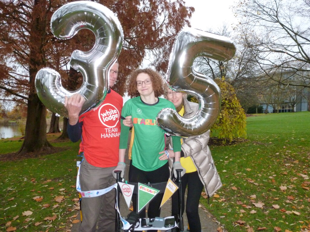 Hannah standing holding on to her four wheeled walker, wearing a green Starlight charity t-shirt, black leggings, splints and purple trainers. She is being supported under her arm by her Dad, Brian, who is wearing a red White Lodge charity t-shirt and holding a silver 3 number balloon. On the other side her Mum, Jill is supporting her under her arm and wearing a yellow Child Bereavement UK charity t-shirt and holding a silver 5 number balloon.