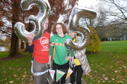 Hannah standing holding on to her four wheeled walker, wearing a green Starlight charity t-shirt, black leggings, splints and purple trainers. She is being supported under her arm by her Dad, Brian, who is wearing a red White Lodge charity t-shirt and holding a silver 3 number balloon. On the other side her Mum, Jill is supporting her under her arm and wearing a yellow Child Bereavement UK charity t-shirt and holding a silver 5 number balloon.