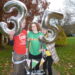 Hannah standing holding on to her four wheeled walker, wearing a green Starlight charity t-shirt, black leggings, splints and purple trainers. She is being supported under her arm by her Dad, Brian, who is wearing a red White Lodge charity t-shirt and holding a silver 3 number balloon. On the other side her Mum, Jill is supporting her under her arm and wearing a yellow Child Bereavement UK charity t-shirt and holding a silver 5 number balloon.