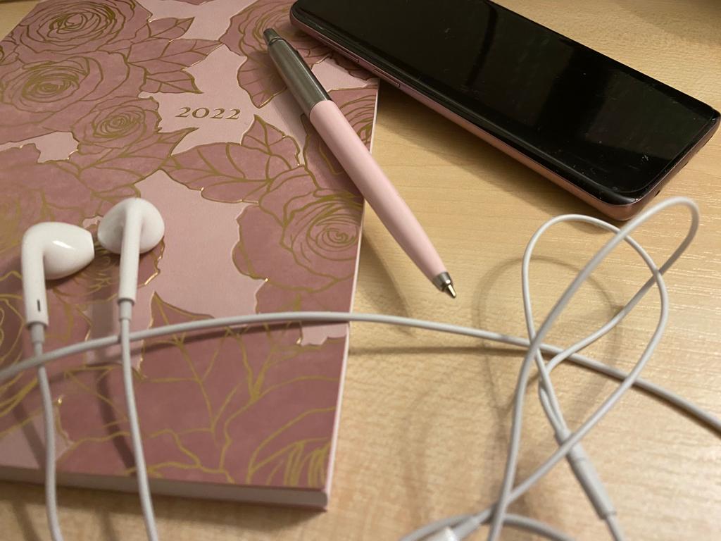A rose gold 2022 diary with gold outline flowers on it. A pink pen, headphones and a phone overlapping it.