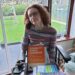 Hannah sitting in her powerchair in the lounge. She is wearing a multi coloured fair isle jumper and has the disability rights handbook and paperwork on her lap. She is talking on the phone, dealing with disability battles and administration.