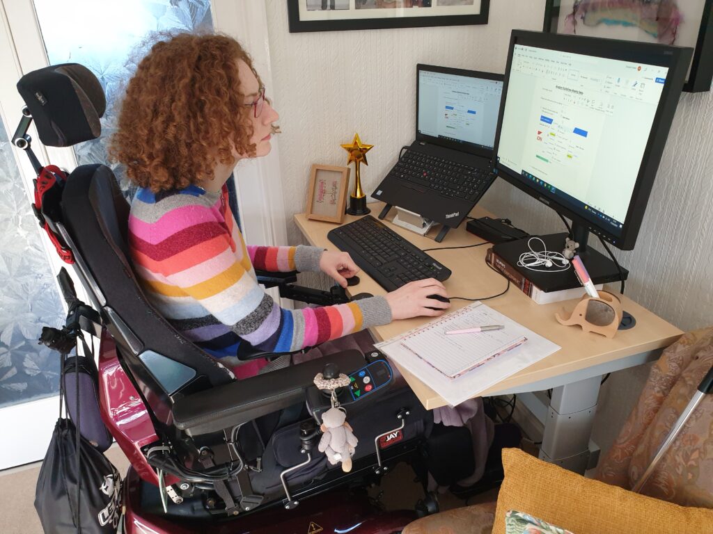 Hannah sitting in her powerchair working at her desk with her laptop and monitor on. She is wearing a multi coloured striped jumper.