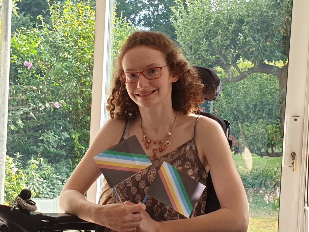 Hannah sitting in her powerchair wearing a brown strappy summer dress with a pale pink pattern and little elephants on it. She is holding two Disability Pride flags in celebration of Disability Pride month and smiling at the camera. The garden is in the background.
