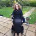 Hannah sitting in her powerchair in her garden, wearing her black RainDek Long Parka, a cover/jacket for a wheelchair user's legs.