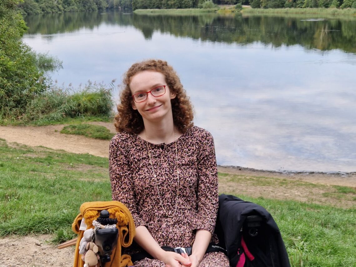 Hannah sitting in her powerchair wearing a black, pink, white and mustard flecky dress over leggings. Behind her is Virgina Water Lakes. The image is for illustrative purposes for stereotypes and the assumptions they make.