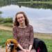 Hannah sitting in her powerchair wearing a black, pink, white and mustard flecky dress over leggings. Behind her is Virgina Water Lakes. The image is for illustrative purposes for stereotypes and the assumptions they make.