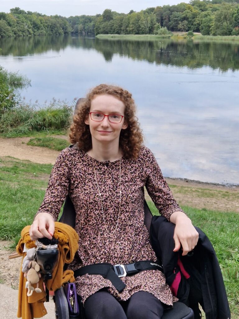 Hannah sitting in her powerchair wearing a black, pink, white and mustard flecky dress over leggings. Behind her is Virgina Water Lakes. The image is for illustrative purposes for stereotypes and the assumptions they make.