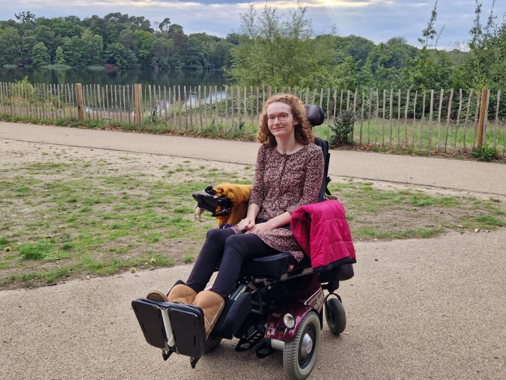 Hannah sitting in her powerchair wearing a black, pink, white and mustard flecky dress over leggings. Behind her is grass and trees and the edge of Virgina Water Lake. The image is for illustrative purposes for stereotypes and the assumptions they make.