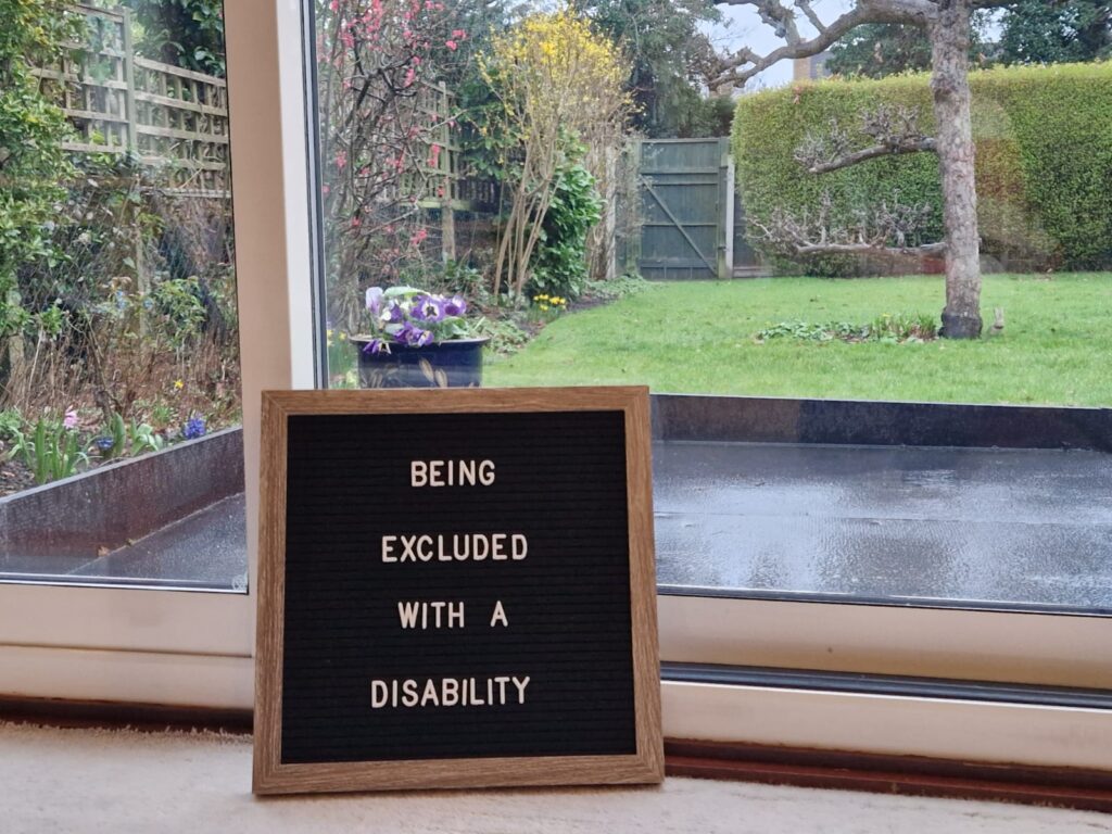 A letter board which is inside the house and says: 'Being Excluded With A Disability' on it. It is leaning against the glass patio door with the garden and outside world beyond it.