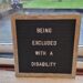 A close up of a letter board which is inside the house and says: 'Being Excluded With A Disability' on it. It is leaning against the glass patio door with the garden and outside world beyond it.