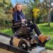 Hannah sitting in her powerchair which has a rose gold base in the garden. She is smiling at the camera and the grass and trees are behind her.