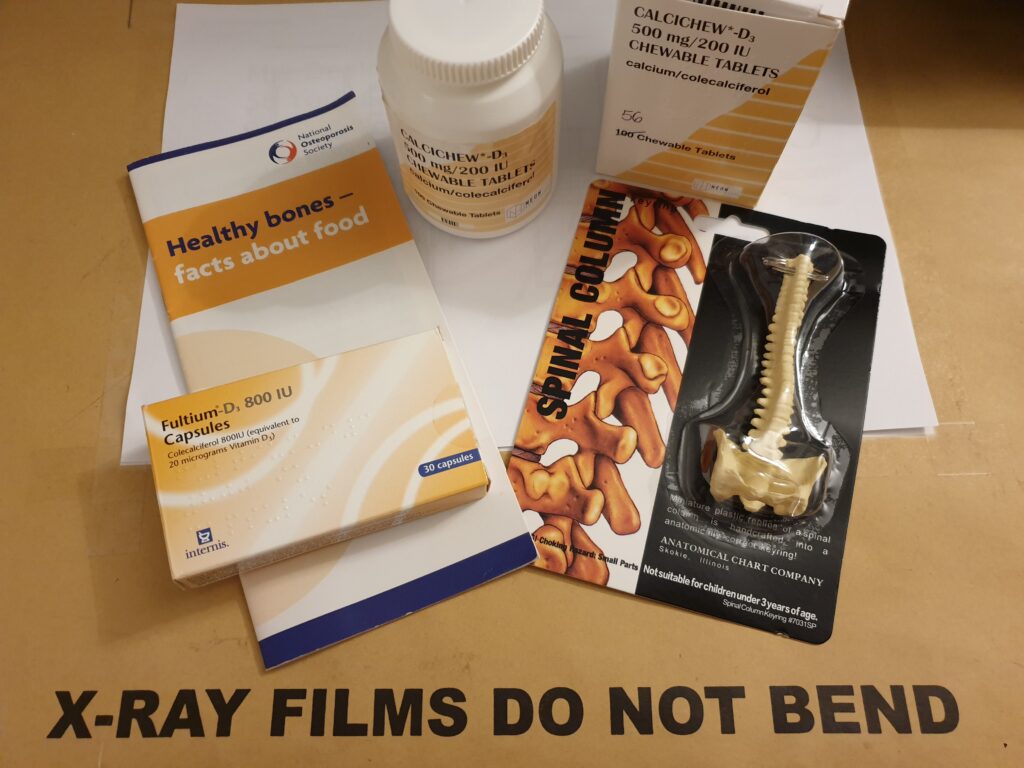 A brown envelope of x-rays which says 'X-ray films do not bend' on the outside. On top of the envelope is a national osteoporosis society healthy bones booklet, vitamin D tablets, Calcichew D3 tablets and a keyring of the spine to illustrate osteoporosis.