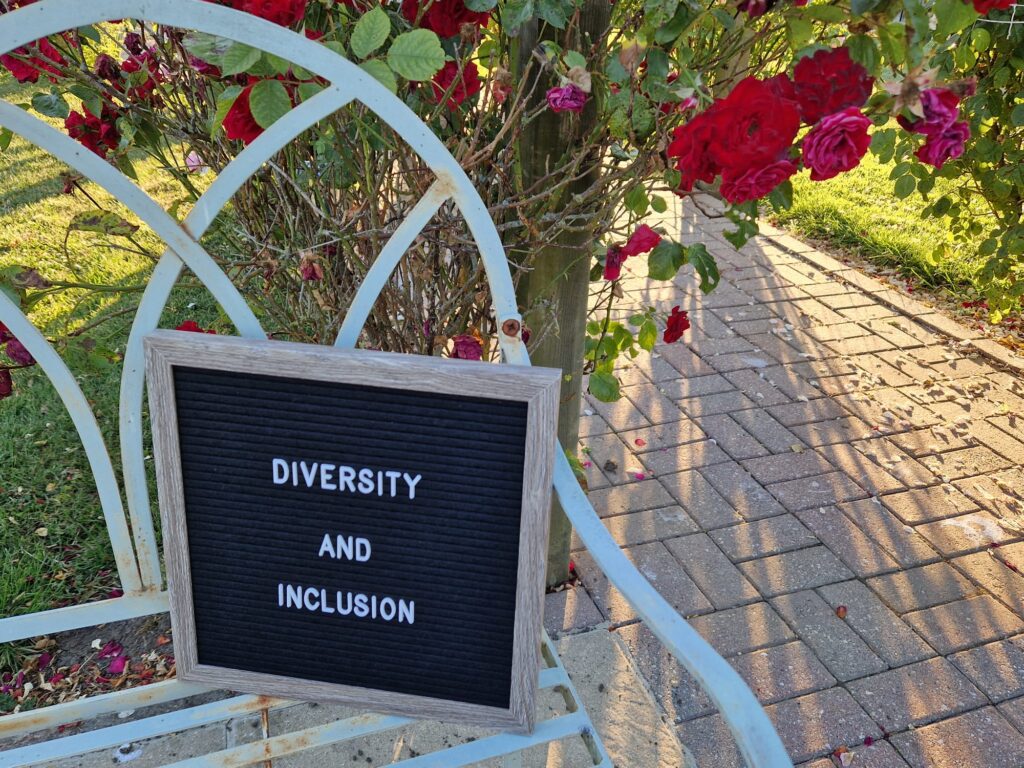 On the left there is a white metal bench which a letter board stands on. The letter board says: 'Diversity and Inclusion'. There are red roses in the background on a pagoda.