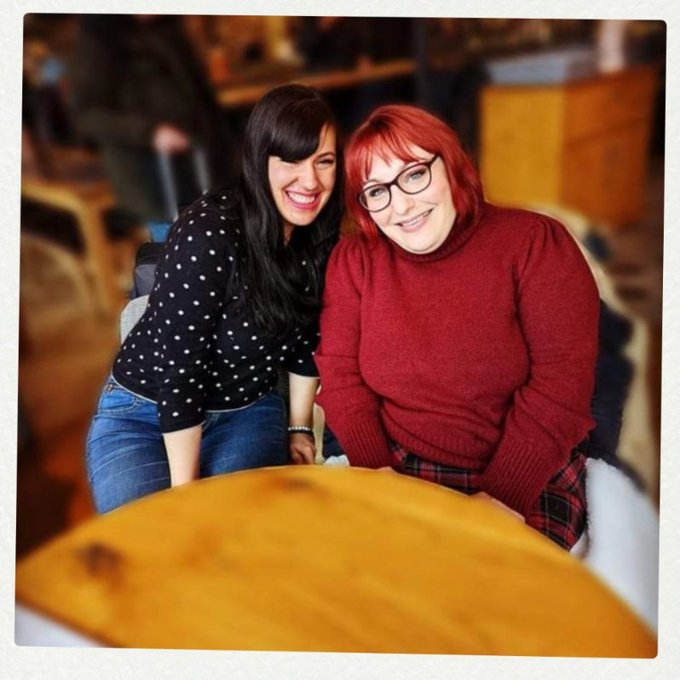 Alice, who has black hair and is wearing a black polka dot top and Lucy who has red hair, glasses and a red jumper are smiling at the camera.