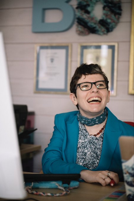 Beth Moulam sitting at her desk laughing. She is wearing a turquoise patterned top, turquoise jacket and necklace, There are framed certificates behind her on the wall.