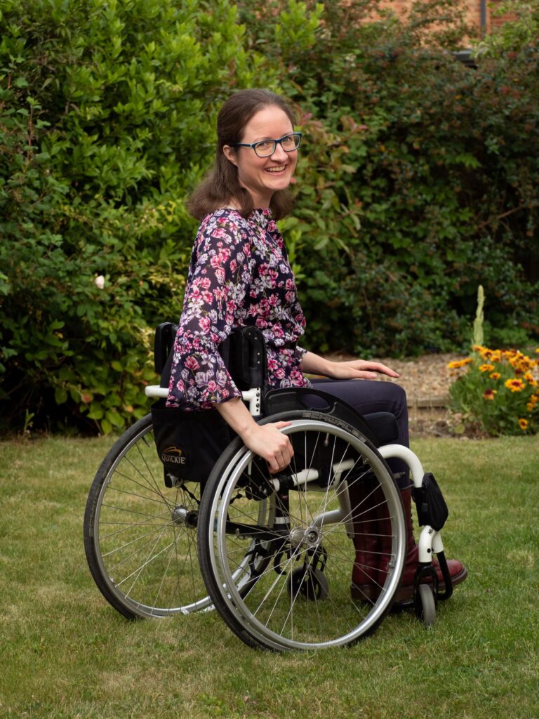 Hannah Ensor, a white woman sitting in her manual wheelchair twisting back to look at the camera. She has black trousers a purple flowery top, long brown hair and glasses. She is on the grass and there is a hedge behind her.