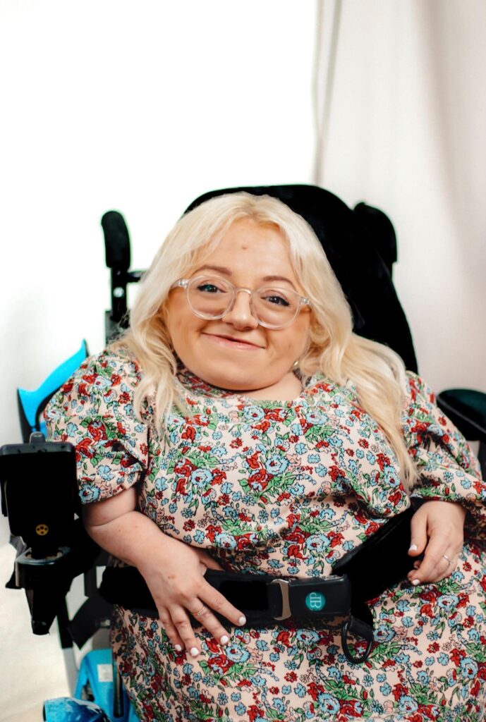 Gem Turner one of the featured disability bloggers sitting in her powerchair wearing a flowery dress. She is a white woman with blonde hair and glasses.