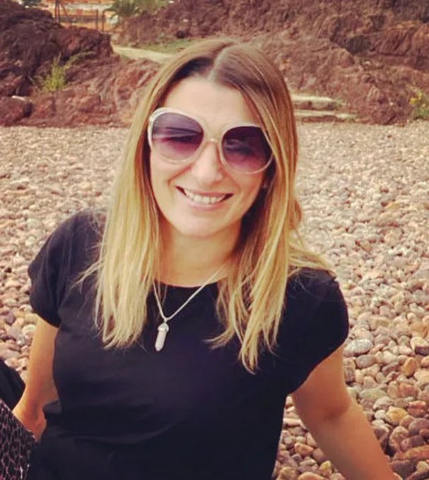 Emma a white woman with blonde hair, sunglassses and smiling at the camera. She is wearing a black t-shirt and sitting on what looks like a stoney beach.