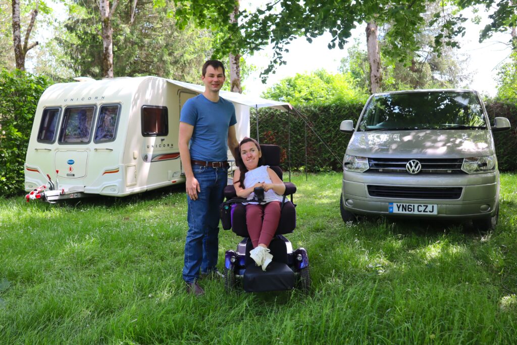 Karla sitting in her powerchair next to Stephen on the grass. Behind them is their caravan and wheelchair accessible vehicle.