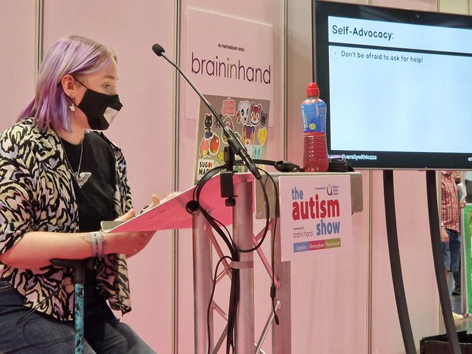 Lozza sitting on a stool in front of a microphone. Lozza is wearing a mask, has purple highlights in her hair, a leopard print blouse and jeans.