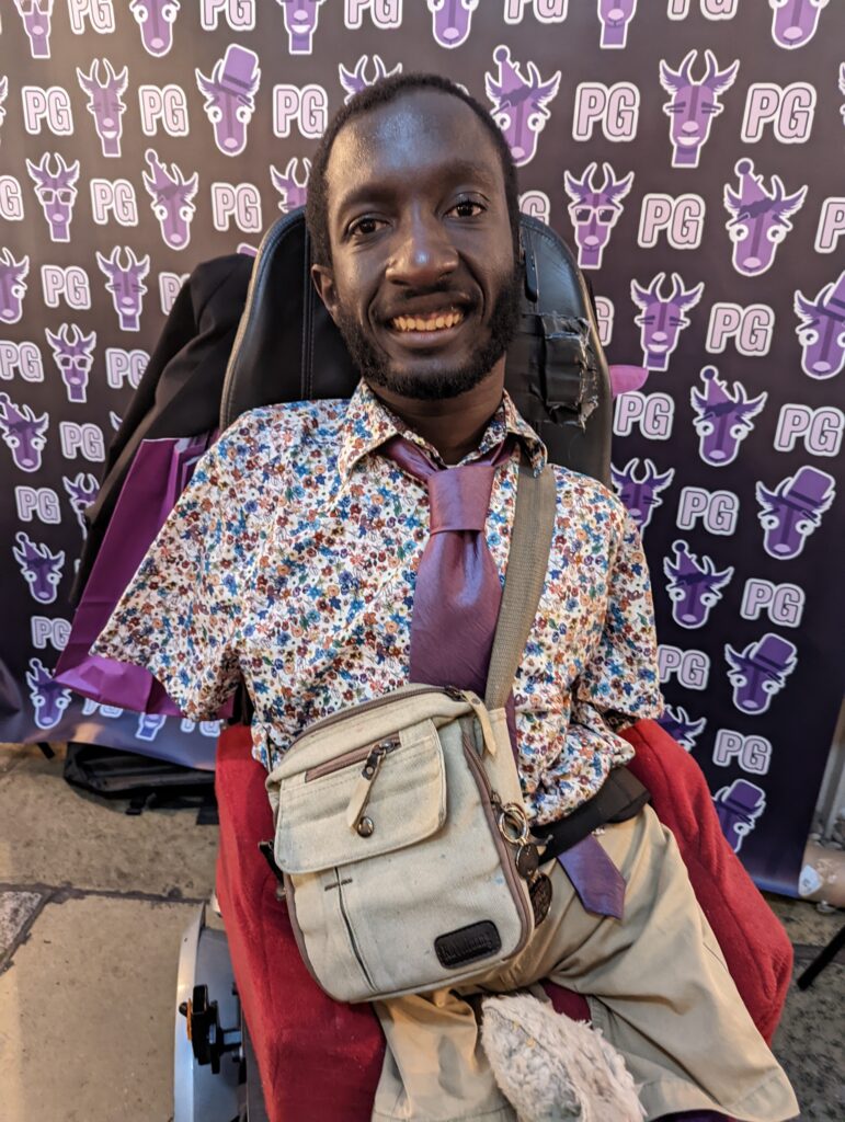 Issac Harvey MBE, a disabled influencer and campaigner. He is a black man sitting in his powerchair, he has a fancy purple patterned shirt, with a purple tie and beige trousers on and is smiling at the camera. Behind him on the wall is the Purple Goat Agency logos,