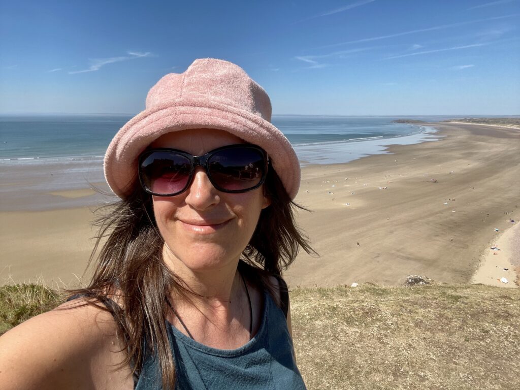 Juliette is wearing a green vest, sunglasses and a pale pink sunhat, she is standing of the beach. You can see the sand and sea in the background.