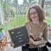 Hannah sitting in her powerchair in the lounge, her garden is in the background. She is wearing a pale pink jumper with pink, white and silver sequin ice skates on it and smiling at the camera. She is holding a letter board which saying '2024- My Hopes and Goals' in white writing.