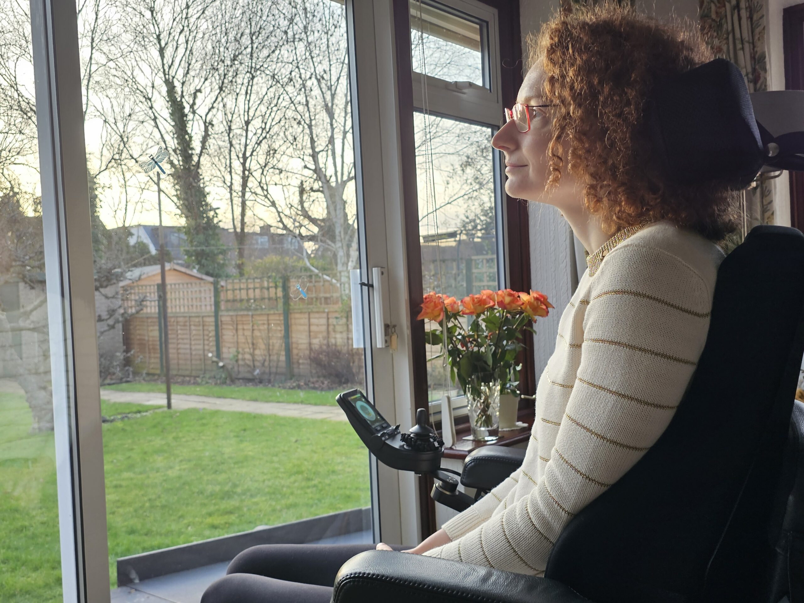 Hannah, a white woman with aburn curly hair and glasses, wearing a cream jumper with gold stripes and leggings and sitting in her powerchair. She is gazing out of the window thinking about the lessons learnt since becoming disabled.
