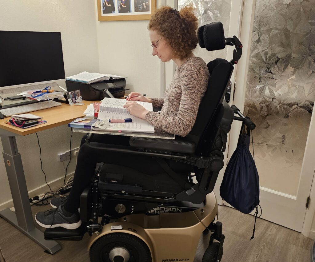 Hannah studying ACCA. She is sitting in her powerchair and has her table that attaches to her powerchair on and thick accounting books on it she is reading.