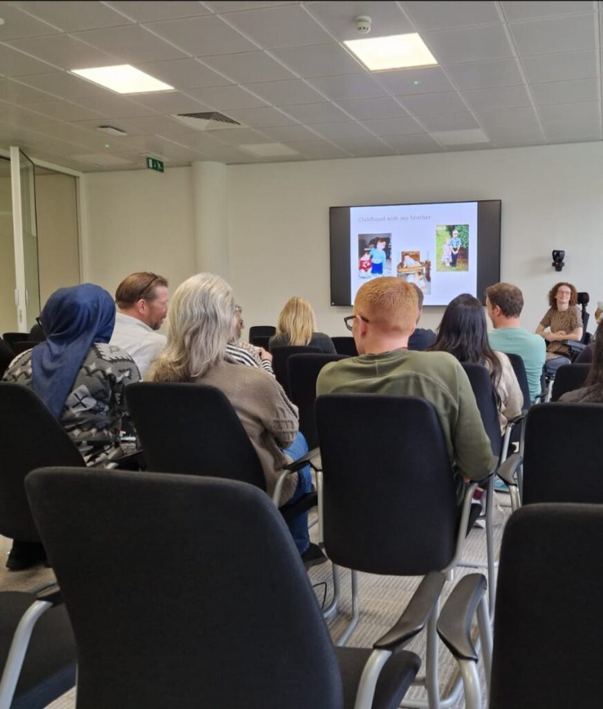 Hannah prior to speaking at work. There is a powerpoint behind her on the wall showing photos of her with her brother. In front of her there are many rows of chairs, slowly filling up with people.