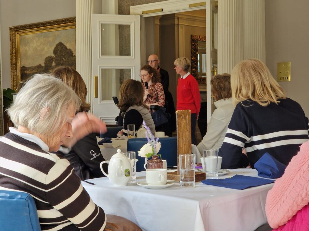 Hannah speaking about White Lodge at Burhill Golf Club. Hannah is in the distance and speaking to a room full of ladies sitting at tables after eating their lunch.
