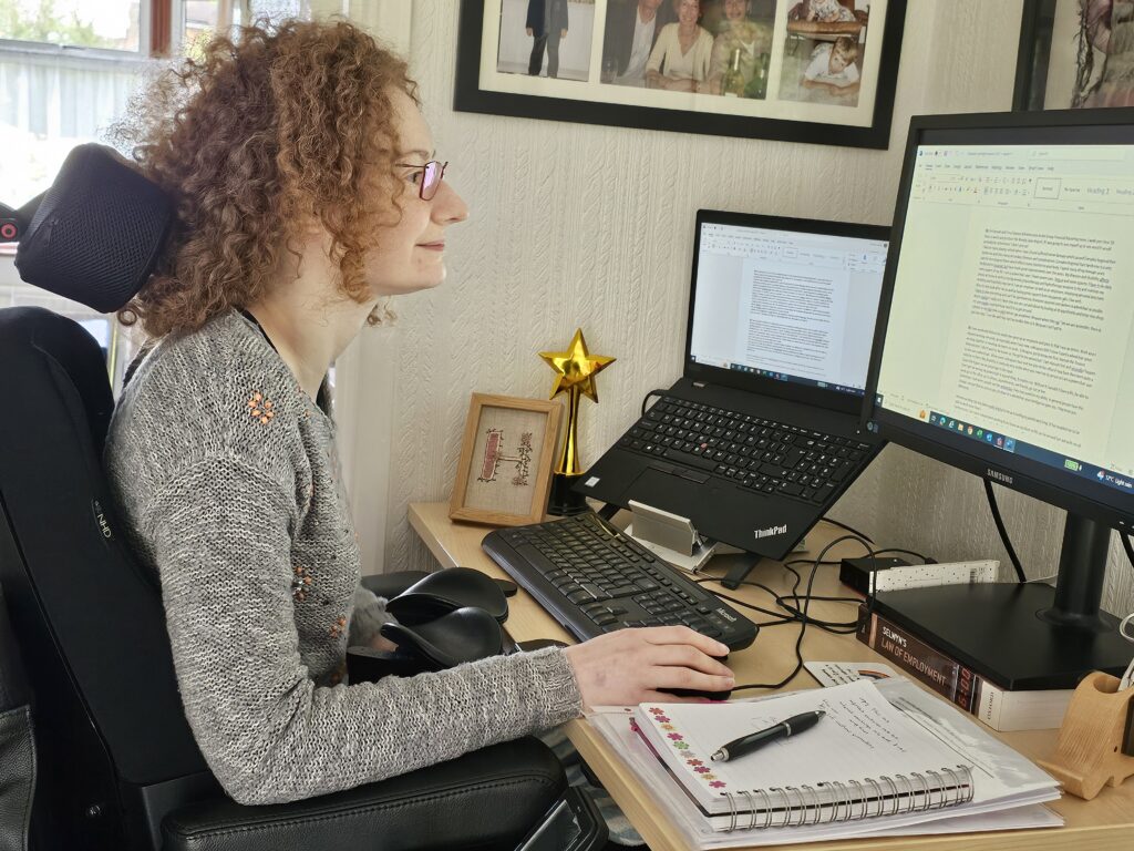 Hannah is working from home, sitting in her powerchair at her desk with her laptop, monitor, keyboard, mouse and notebook on. She is focusing on the computer screen.